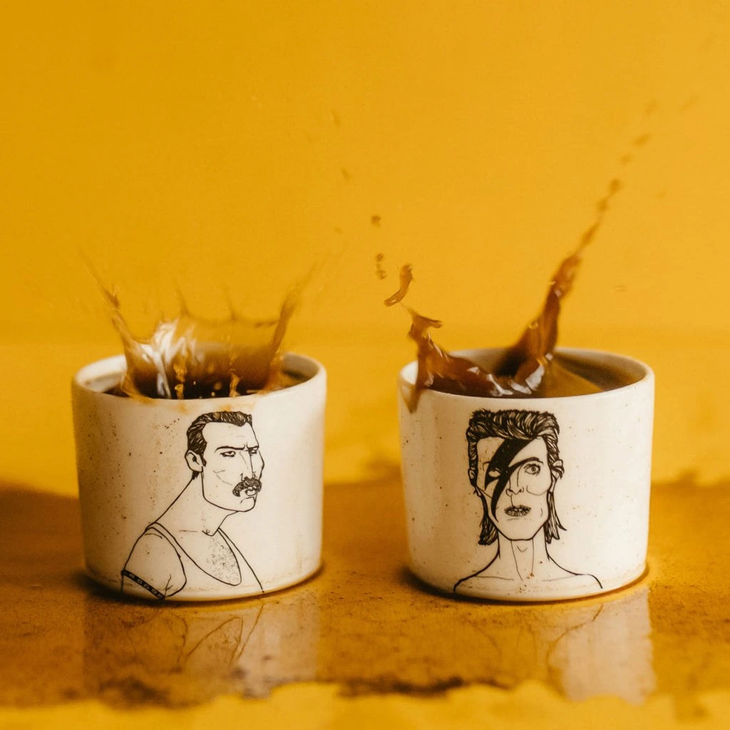 The Legends Espresso set features two ceramic mugs with illustrated faces—a moustache and lightning bolt—splashing coffee on a yellow backdrop, perfect for Tallinn shopping or unique Estonian design inspiration. Gate21.ee
