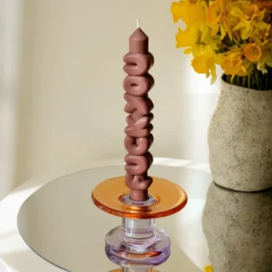 The F*ck You 3D Candle - Mocha stands upright in a glass holder with orange and purple accents on a mirrored surface, making it ideal for a concept shop or gift shop. A vase with yellow flowers appears in the background. Gate21.ee