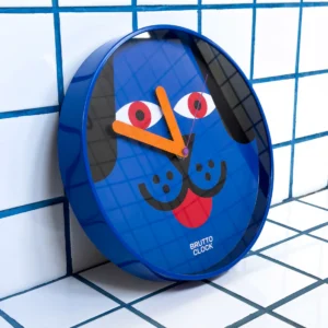 The DOGGO - Wall Clock features a blue frame, orange hands, and a dog face design with eyes, nose, and tongue. "BRUTTO CLOCK" is printed on it. The clock highlights unique Estonian design against white tiles with blue grout lines. Gate21.ee