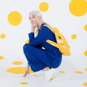 A person with long, fair hair in a blue outfit and white trainers crouches on a floor of large yellow polka dots, carrying the Golden Dots - Notabag—a yellow bag with white spots—against a matching spotted white wall and floor. Gate21.ee