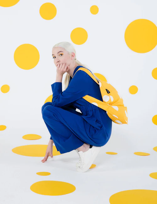 A person with long, fair hair in a blue outfit and white trainers crouches on a floor of large yellow polka dots, carrying the Golden Dots - Notabag—a yellow bag with white spots—against a matching spotted white wall and floor. Gate21.ee