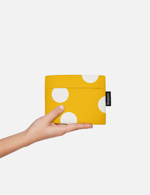 A hand holds the Golden Dots - Notabag, a small yellow pouch with bold white polka dots and a distinct black Baggu tag, ideal for any curated gift shop, set against a plain white background. Gate21.ee