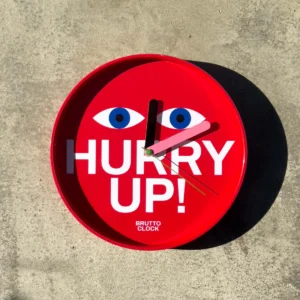 The HURRY UP! - Wall clock features red casing, bold white “HURRY UP!” text, blue and white cartoon eyes, black hour and minute hands, and a red second hand—an eye-catching piece of Estonian design on a concrete surface. Gate21.ee