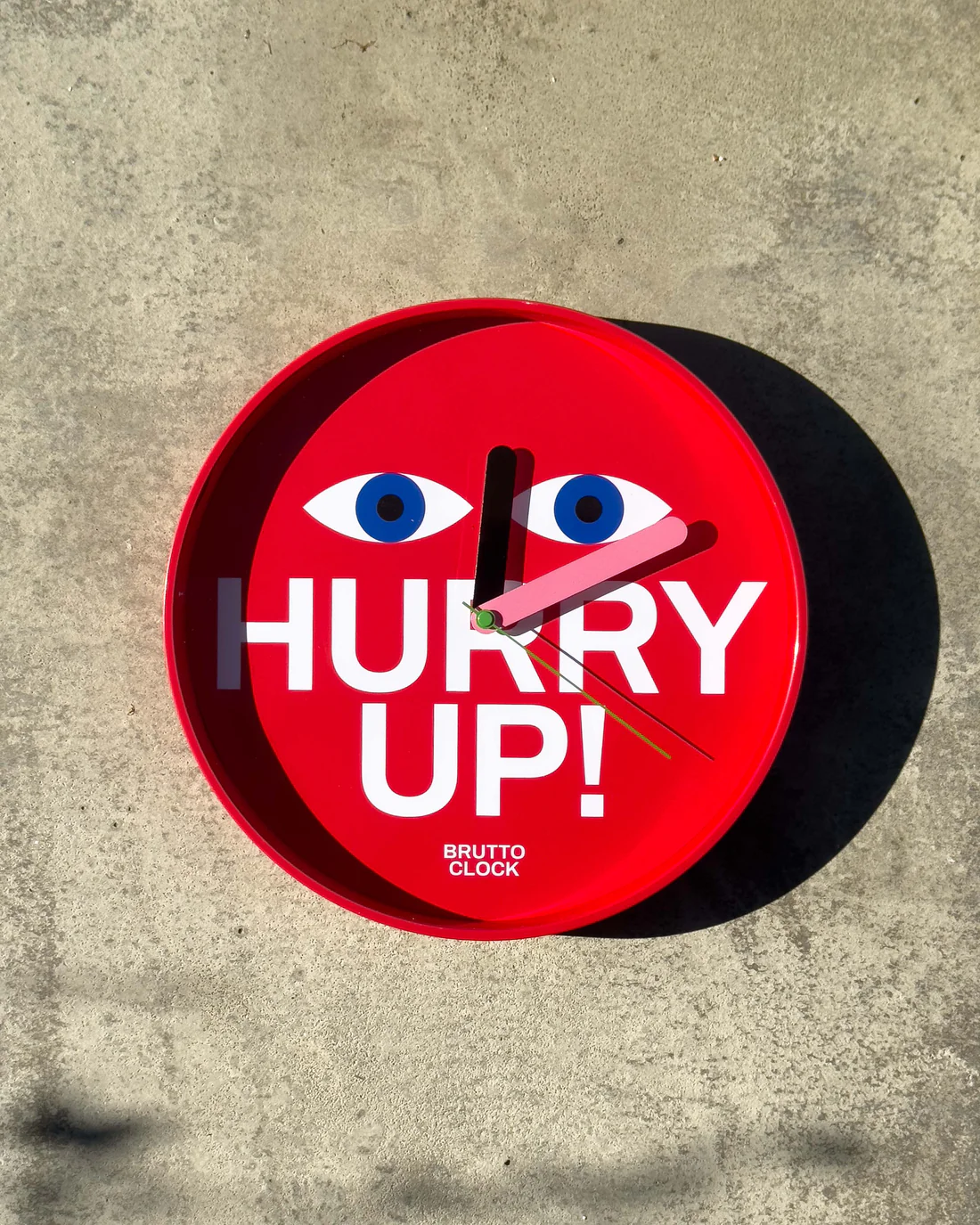 The HURRY UP! - Wall clock features red casing, bold white “HURRY UP!” text, blue and white cartoon eyes, black hour and minute hands, and a red second hand—an eye-catching piece of Estonian design on a concrete surface. Gate21.ee