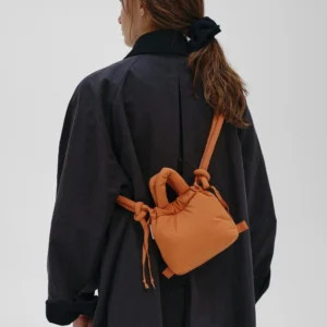 A person with long brown hair tied back, in a dark coat and carrying the Micro Ona Soft Bag - Oxide with knotted straps, stands facing away from the camera against a plain light background. Gate21.ee