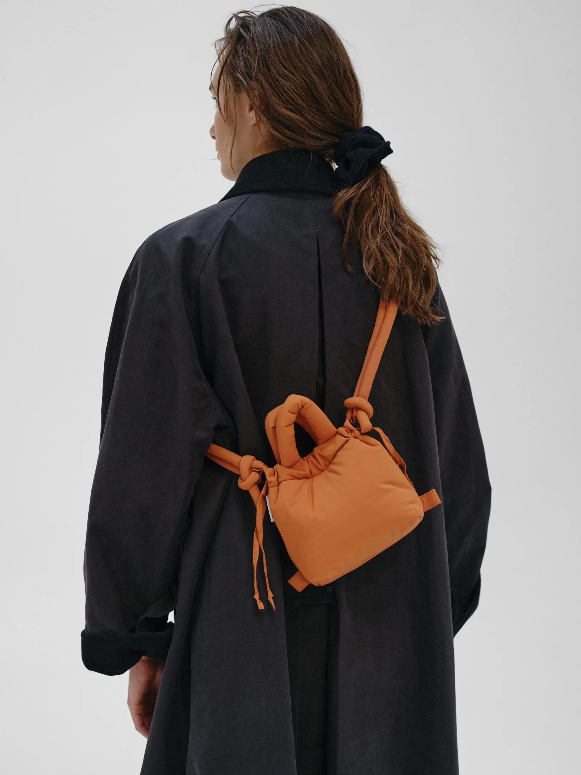 A person with long brown hair tied back, in a dark coat and carrying the Micro Ona Soft Bag - Oxide with knotted straps, stands facing away from the camera against a plain light background. Gate21.ee