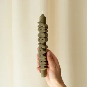 A hand holds the Happy Bday 3D Candle in olive green, featuring raised "HAPPY BDAY" letters wrapped around it. Set against a plain light curtain, it's perfect for unique celebrations or gifting from a concept shop. Gate21.ee