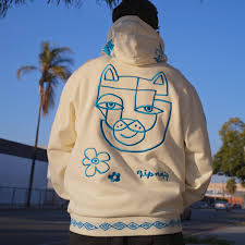 Wearing the Blonded hoodie - Off White, featuring a bold blue abstract cat face, flower, and geometric patterns on the back, someone stands outside with trees and a building in the background. Gate21.ee