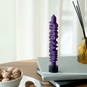 The Happy Bday 3D Candle - Indigo, an Estonian-designed tall purple candle with "Happy Bday" moulded vertically, stands on a black holder atop grey books beside a yellow reed diffuser and a bowl of walnuts—perfect for unique gift ideas. Gate21.ee