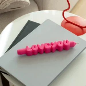 The Love You 3D Candle - Pink sits on a closed grey laptop and black notebook on a round white table, making it a charming gift or desk accent. A red lamp and beige sofa are visible in the background. Gate21.ee