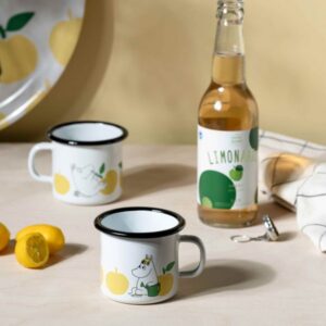 A light wooden surface displays a bottle of lemonade, sliced lemons, a patterned tray, and the Joyful Apples – Moomin enamel mug 2.5dl—perfect inspiration for Tallinn shopping or a find from your favourite gift shop. Gate21.ee