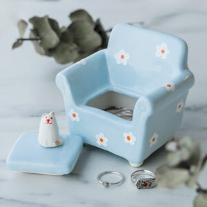The Blue Chair - Jewellery Box is a light blue ceramic armchair with white flower patterns and a removable lid topped by a cat figurine. Displayed on marble with two rings nearby, it’s an ideal gift for lovers of unique Estonian design. Leaves add a natural touch. Gate21.ee