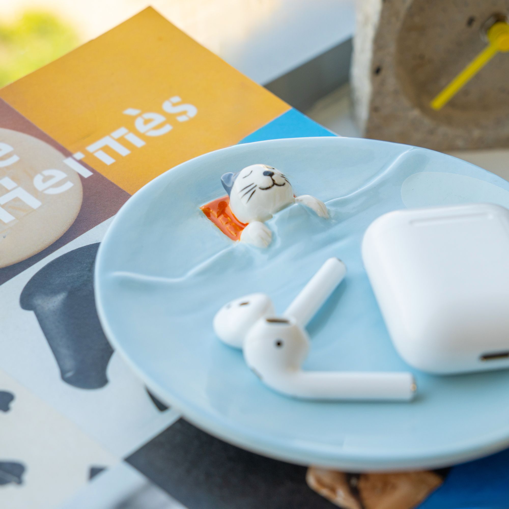 The Blue Dreamer Cat - Jewellery Plate is a small ceramic dish shaped like a pool with a cat figurine lounging on an orange lilo—an adorable Estonian design ideal for your Tallinn shopping finds. A colourful booklet and a concrete clock are nearby. Gate21.ee