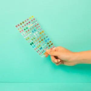A hand holds a clear sheet of Kawaii - Earring Stickers with small, colourful curated designs, set against a solid turquoise background. Gate21.ee