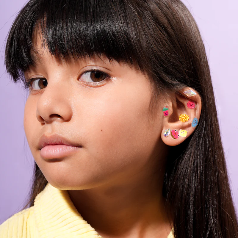 A young girl with long dark hair and fringe looks at the camera, wearing a pale yellow top and Kawaii - Earring Stickers on her cheek and ear, set against a solid light purple background that evokes Tallinn’s creative gift shop vibe. Gate21.ee