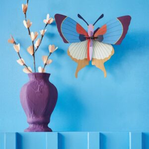 A Plum Fringe Butterfly decorates the wall above a purple vase with beige flower branches on a blue shelf, creating a whimsical display in a charming Tallinn gift shop. Gate21.ee