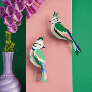 Forest Crests: Two green and white paper birds with blue accents, inspired by Estonian design, are attached to a vertical pink panel beside a purple vase holding pink foxgloves, all set against a green background. Gate21.ee