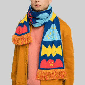 A person wears Moodies – Organic: a mustard yellow jacket, pink shirt, and a blue scarf with vibrant cartoon patterns, standing before a light grey background—perfect for showcasing in a curated gift shop or concept store. Gate21.ee