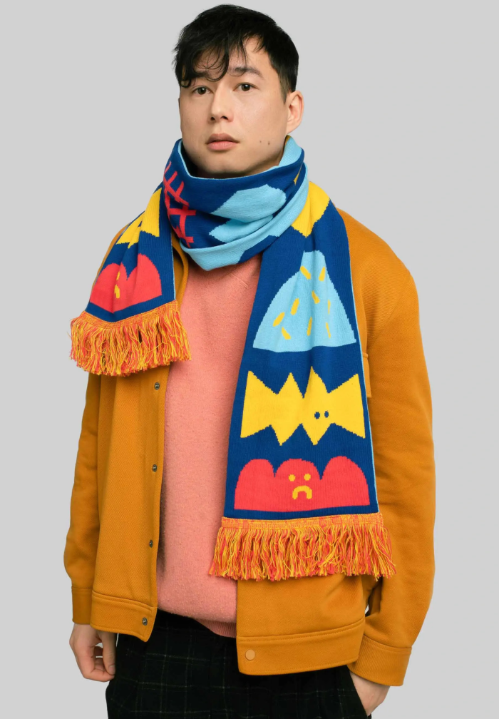 A person wears Moodies – Organic: a mustard yellow jacket, pink shirt, and a blue scarf with vibrant cartoon patterns, standing before a light grey background—perfect for showcasing in a curated gift shop or concept store. Gate21.ee