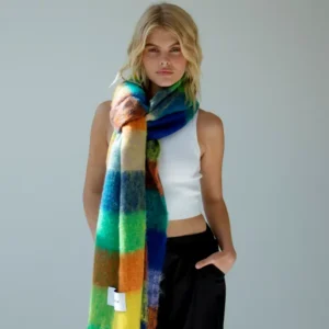 A person with shoulder-length blonde hair wears a white sleeveless top, black trousers, and The Reykjavik Scarf - Nordic Skies. One hand is in their pocket, with a plain light grey background. Gate21.ee