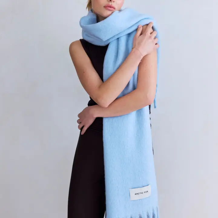 A person in a sleeveless black outfit is wrapped in The Stockholm Scarf - Glacier Blue, which is large, light blue, and features fringe at the ends. A white Arctic Fox label is visible on the scarf against a plain, light-coloured background. Gate21.ee