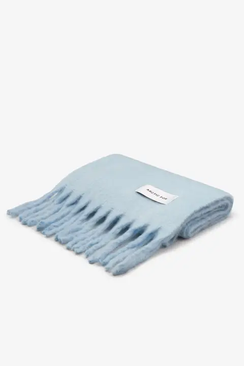 The Stockholm Scarf - Glacier Blue, a light blue wool scarf with fringed edges, is neatly folded and features a white rectangular Arctic Fox Stockholm label sewn onto its surface. Gate21.ee