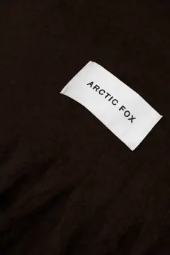 A white rectangular fabric label with "ARCTIC FOX" in black capitals is sewn onto The Reykjavik Scarf - Ground Coffee, a dark brown, textured accessory. Gate21.ee
