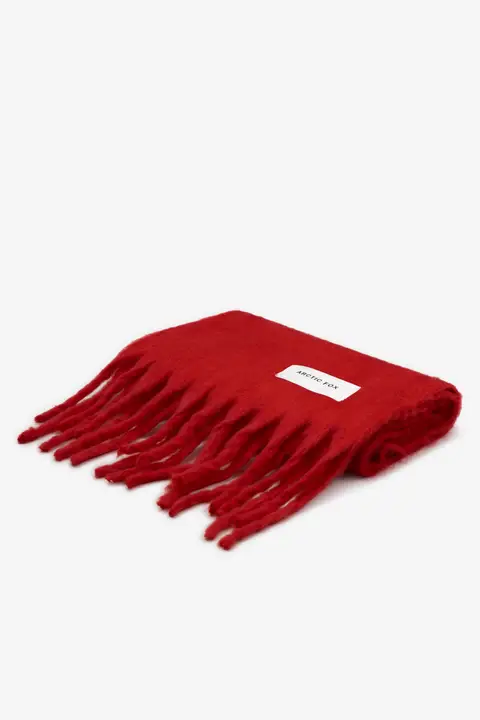 The Stockholm Scarf - Crimson Red from Arctic Fox is a bright red wool scarf with long fringe on one end and a rectangular white label sewn onto the visible corner, neatly folded against a plain white background. Gate21.ee