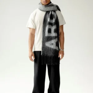 A person faces forwards in a white T-shirt, black pleated trousers, grey and white trainers, and the Arctic Fox Scarf - Monochrome Logo featuring bold lettering. The background is plain and light-coloured. Gate21.ee