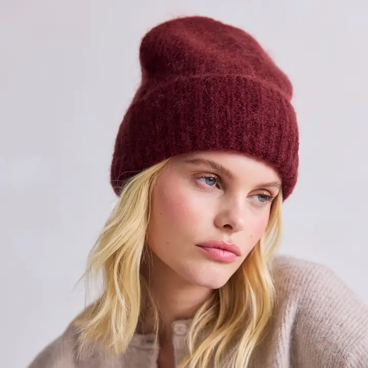 A person with long blonde hair wears the Mohair Beanie - Cherry Burgundy and a beige jumper, gazing slightly to the side against a plain light background. Gate21.ee