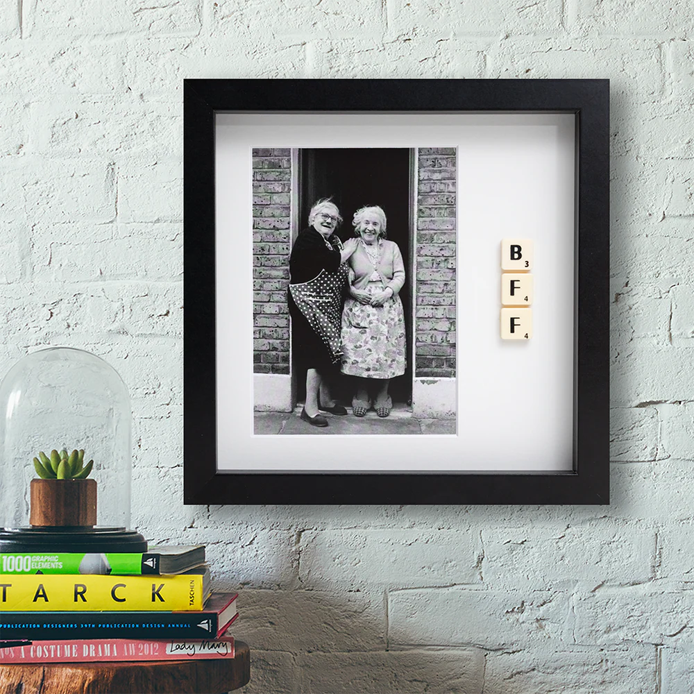The BI POLAR - Scrabble Print features a framed black-and-white photo of two smiling elderly women with “BFF” spelt out in Scrabble tiles, displayed stylishly on a white brick wall above books for a charming touch. Gate21.ee