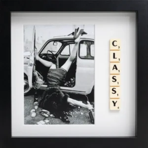 The CLASSY - Scrabble Print features a black and white photo of someone in a dress climbing into a small car, legs raised, amid bottles. Scrabble tiles spelling “CLASSY” sit nearby—making this playful print perfect for sparking fond memories. Gate21.ee