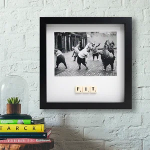 A framed black-and-white photo titled "FIT - Scrabble Print" hangs on a white brick wall, with Scrabble tiles spelling “F.I.T.” above stacked books and a glass dome housing a small plant. Gate21.ee