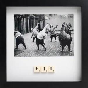 The FIT - Scrabble Print features a black and white photo of women in jumpers and skirts stretching together in a courtyard, with "FIT" spelt out in Scrabble tiles beneath the image, all set in a sleek black frame. Gate21.ee