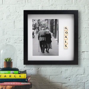 The GOALS - Scrabble Print, a framed black-and-white photo of an elderly couple walking arm in arm with Scrabble tiles spelling "GOALS," is displayed on a white wall beside a stack of books and a plant—perfect for sparking cherished memories. Gate21.ee