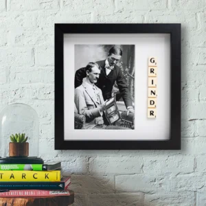 A GRINDR - Scrabble Print sits on a shelf beside a framed black-and-white photo of two men, with books and a glass dome holding a small plant below—perfect for sparking memories or celebrating moments that matter. Gate21.ee