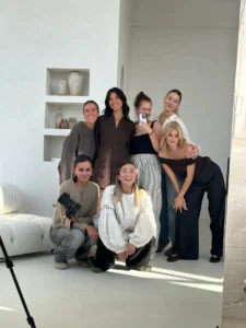 Six women pose in a bright, minimalist room&mdash;some standing, some kneeling. One holds a camera while another takes a photo with her phone in the mirror. The Reykjavik Scarf - Nordic Skies is worn, with shelves and vases visible in the background. Gate21.ee