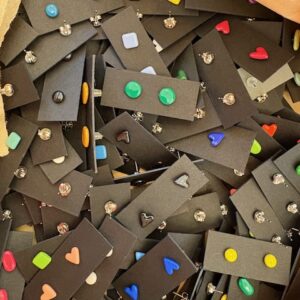 A pile of ABE jewellery - Small shapes - K&Otilde;IK black rectangular cards, each showing a pair of colourful stud earrings in assorted heart, circle, and square shapes, arranged randomly. Gate21.ee