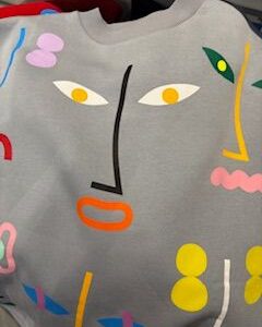 A grey fabric displays a colourful abstract face design, featuring geometric shapes in yellow, orange, black, white, pink, green, and blue for the facial features and accents. Gate21.ee