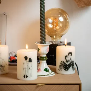 A Back - Candle with black line drawings of couples sits alongside a twisted green candle in a floral holder and a Helen B ceramic soap dish; all rest on wood whilst a large exposed bulb lamp glows behind. Gate21.ee