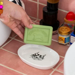 A hand holds a green "Schafmilchseife" sheep soap above the Kitty - Tray, a Helen B ceramic soap dish with a black cat face, set on pink tiles next to toiletries. Gate21.ee
