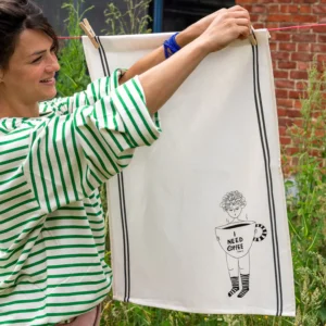 A person hangs the Couple Tea Towel - Need Coffee, featuring Helen B’s black illustration of a figure with a large coffee mug, on an outdoor washing line while wearing a green and white striped shirt. Gate21.ee