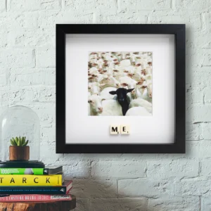 The ME - Scrabble Print features a framed photo of a black sheep among white sheep on a white brick wall. Below, Scrabble tiles spell "ME," accented by books and a plant under glass on the shelf. Gate21.ee