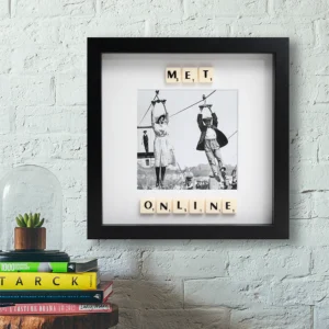 The MET ONLINE - Scrabble Print features a framed photo of two people ziplining, with Scrabble tiles spelling “MET ONLINE,” displayed on a white brick wall above a table with books and a glass dome, evoking cherished memories. Gate21.ee