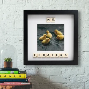 The NO FLOATERS - Scrabble Print features five yellow ducklings swimming, with Scrabble tiles spelling “NO, FLOATERS.” This framed photo adds charm above books or plants—perfect for sparking memories or capturing special moments. Gate21.ee