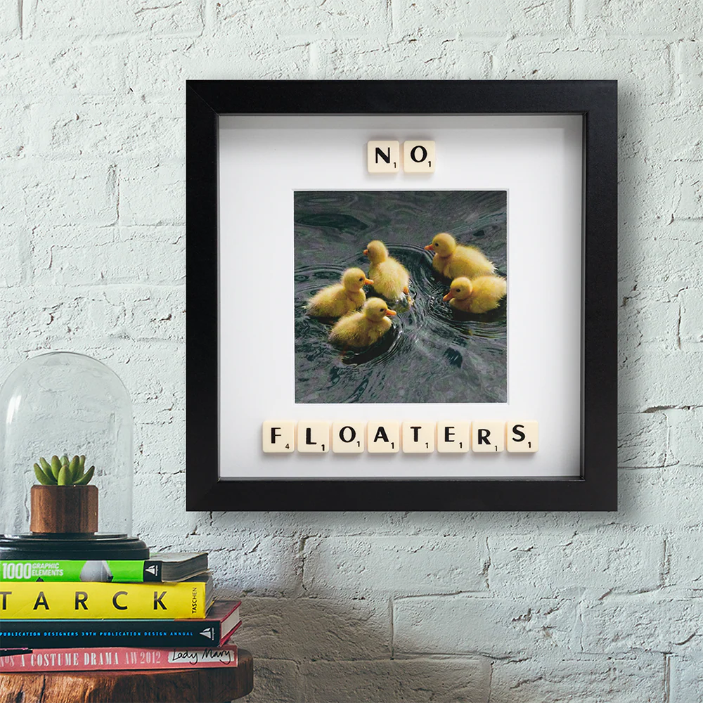 The NO FLOATERS - Scrabble Print features five yellow ducklings swimming, with Scrabble tiles spelling “NO, FLOATERS.” This framed photo adds charm above books or plants—perfect for sparking memories or capturing special moments. Gate21.ee