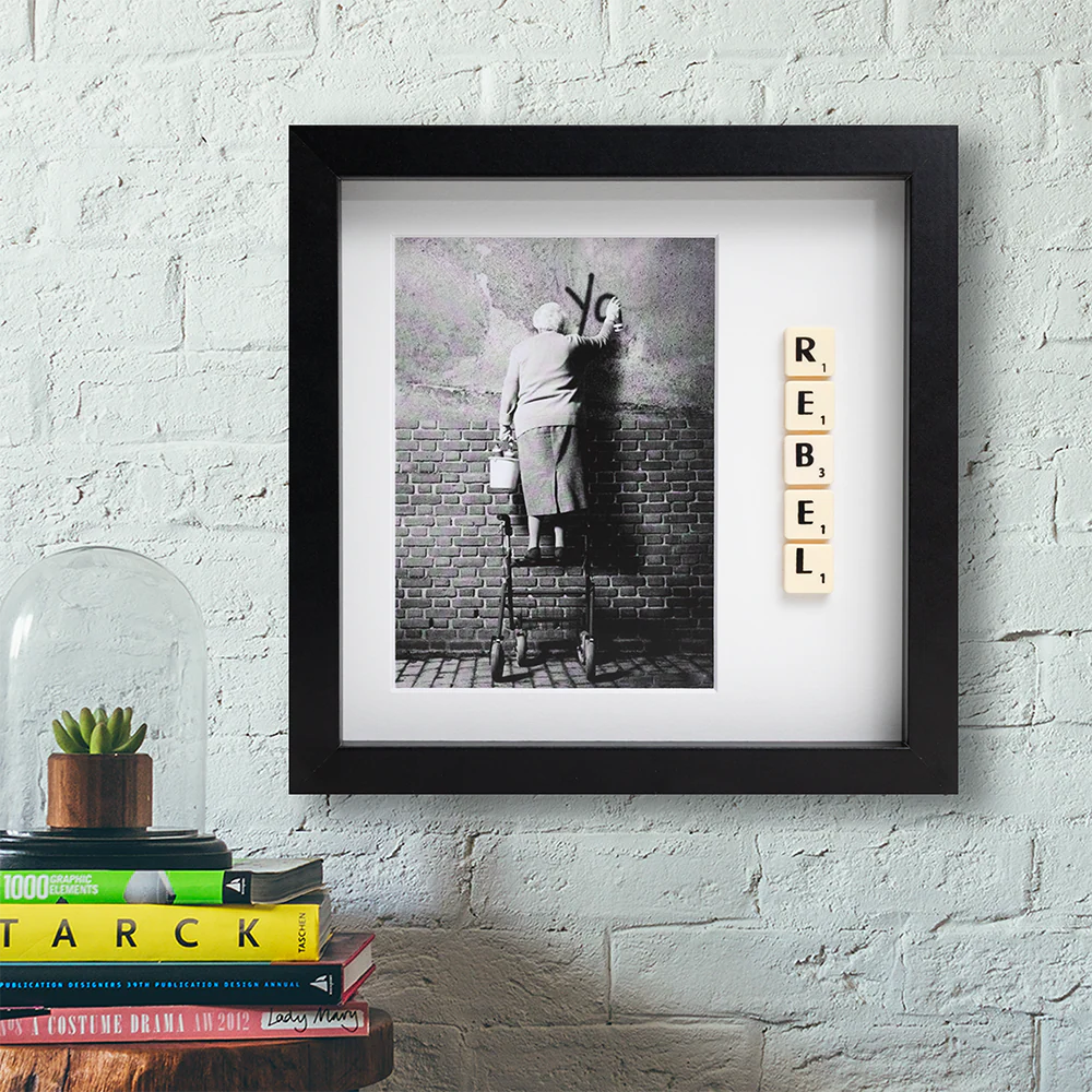 The REBEL - Scrabble Print is a framed black-and-white photo of someone painting graffiti on a brick wall, featuring Scrabble tiles spelling "REBEL." Displayed above books and a glass dome with a plant, it's perfect for sparking memorable moments. Gate21.ee