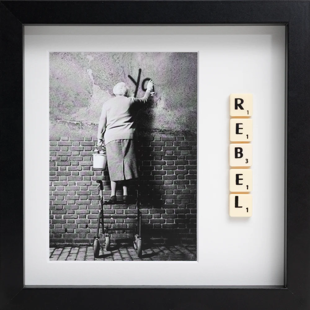 The REBEL - Scrabble Print features a black-and-white photo of an elderly person with a walking frame painting graffiti, with Scrabble tiles spelling “REBEL” beside it. This striking image comes framed in black. Gate21.ee