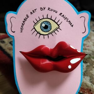 Riivo 3D small brooch features a flat, cartoon face on a pink background with one blue eye, a yellow crown, and large red 3D lips. Above the eye is the text: &ldquo;Wearable Art by Rino Raifstma.&rdquo;. Gate21.ee