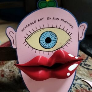 The Riivo 3D suur pross features a flat paper face with a large blue eye, cartoon ears, yellow tuft of hair, and big red lips. &ldquo;WEARABLE ART BY RIINU RAIESMAA&rdquo; appears above the eye. The brooch is displayed on a multicoloured carpet. Gate21.ee